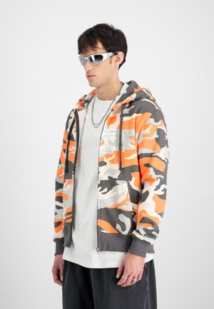Young man wearing silver sunglasses, orange-gray-white camo zip-up hoodie over white shirt and black pants against plain light background.