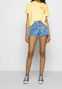 Nainen, jolla on päällään keltainen Levi's T-paita, siniset korkeavyötäröiset farkkushortsit, joissa on suurikokoinen kirjapainoteksti, ja mustat Converse-kengät, seisoo valkoista taustaa vasten.