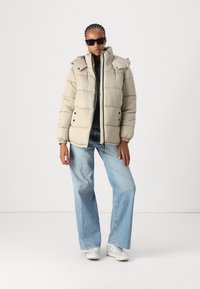 Beige puffjakke med høy krage, glidelåslukking og elastiske mansjetter. Båret over en mørk topp, kombinert med lyseblå flared jeans og sneakers.