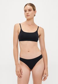Woman wearing a black bralette and matching bikini-style underwear, standing against a plain white background.