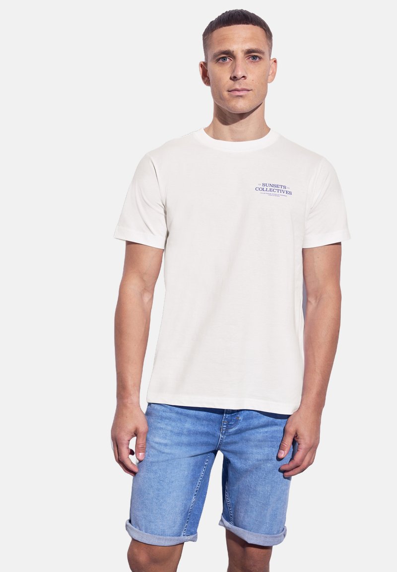 Bílý bavlněný tričko s krátkými rukávy a kulatým výstřihem, s modrou tištěnou logem na hrudi. Nosené s džínovými šortkami světle modré barvy.