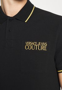 Svart polo-shirt med guldtrim på kragen och ärmarna, med guldbroderad "VERSACE JEANS COUTURE" text på bröstfickan.