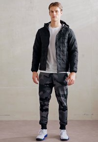 Giacca nera trapuntata con cappuccio, t-shirt bianca sotto e pantaloni jogger grigi camouflage. Completata con scarpe da ginnastica bianche e dettagli neri.