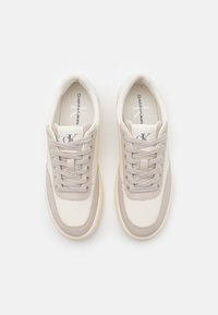 Tenisky Calvin Klein v krémové a světle béžové barvě s koženými a textilními panely, plochými tkaničkami a logem na jazyku. Zaoblený design špičky.