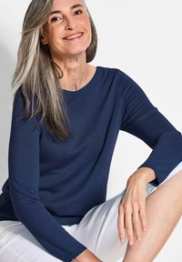 Dunkelblaues, geripptes Langarmshirt mit Rundhalsausschnitt, kombiniert mit weißen Shorts. Das Model hat glattes, langes graues Haar.