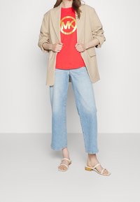 Woman wearing a beige blazer, red t-shirt with gold MK logo, light blue jeans, and beige open-toe sandals with low heels.