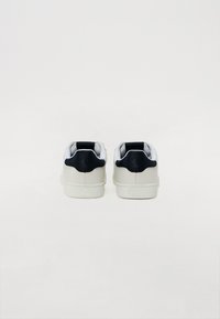 Hvite sneakers med overdel i glatt lær, svart hælakkent, rund tå og gummisåle. Har et minimalistisk design uten mønstre.