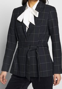Granatowa marynarka w kratę, z paskiem w talii, szpiczastymi klapami i białym kołnierzykiem z muszką. Teksturowana tkanina ze złotymi akcentami.