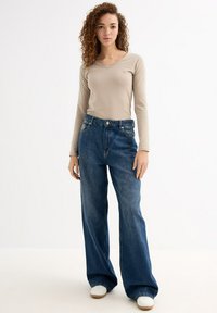 Camisa de manga larga beige con escote en V combinada con unos vaqueros de denim azul de tiro alto y corte holgado. Zapatillas blancas completan el atuendo.