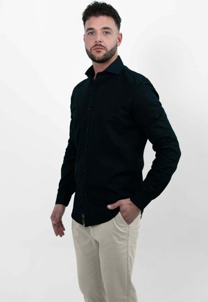 Man met kort donker haar en baard, gekleed in een zwart overhemd met knopen en lichtbeige broek, staand met één hand in de zak tegen een witte achtergrond.