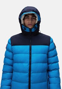 Giacca a vento blu con pannello superiore blu navy, chiusura con zip, cappuccio regolabile con cordino e texture trapuntata. Patches decorativa sulla spalla sinistra.