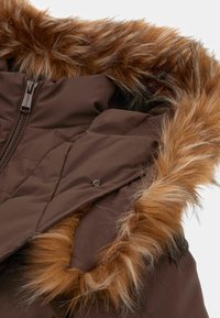 Rudas izoliotas džemperis su dirbtinio kailio kapuliu. Savybės: užtrauktukas ir sagų detalės, pabrėžiančios minkštas tekstūras ir šiltą dizainą.