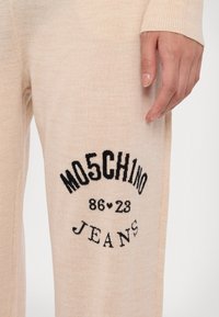 Bež pletene hlače na žabe z črnim grafičnim potiskom, ki vsebuje besedi »MO5CH1NO« in »JEANS« ter leto »86♥23« v središču. Mehka tekstura.