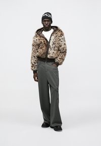 Fuskpälsjacka med leopardmönster och bruna detaljer, kombinerad med gråa vida byxor med kritstrecksrand. Svart mössa med vitt grafiskt mönster.