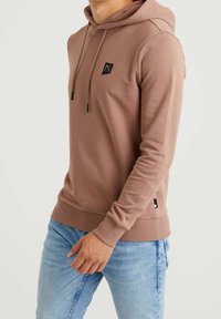Hnědá mikina s texturovaným povrchem, přední kapsou, stažitelnou kapucí a malým černým logem na hrudi. V kombinaci se světle modrými džíny.