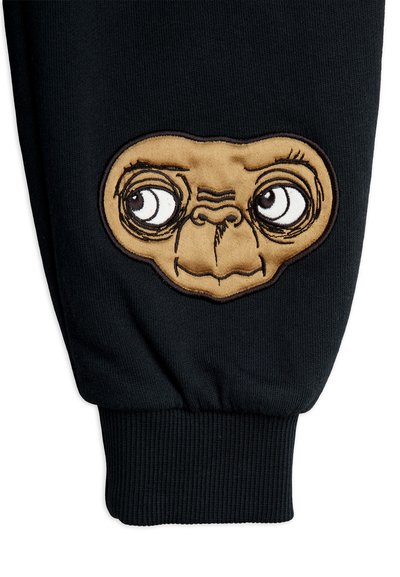 Czarne spodnie z wyraźną brązową wyszywaną łatą przedstawiającą twarz kreskówkowego obcego, z wyraźnymi oczami i teksturowanymi detalami.