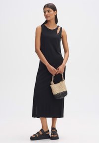 Zwarte mouwloze maxi-jurk met een ronde halslijn en een uitsnijding, gecombineerd met een rieten tas en zwarte platte sandalen.