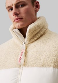 Kremowa polarowa kurtka z wysokim kołnierzem, z białą dolną częścią i czerwoną metką z logo Calvin Klein na zamku.