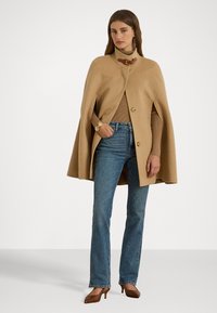 Κάπα από καμήλα με ψηλό γιακά, δύο μπροστινά κουμπιά και πλαϊνές σχισμές, συνδυασμένη με καφέ ριγέ πουλόβερ και μπλε καμπάνα τζιν.