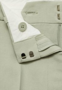 Close-up van de tailleband van een beige broek met haak-en-oog sluitingen, een knoop, een rits en een witte binnenvoering.