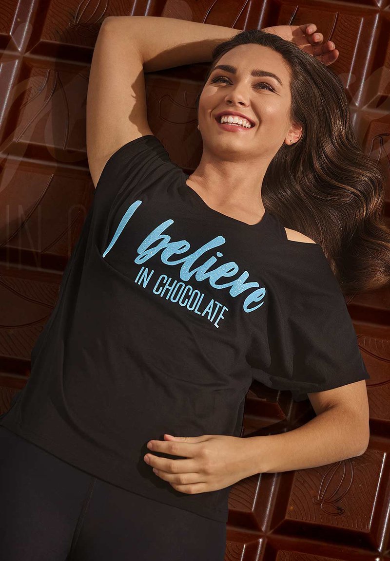 Black t-shirt with "I believe in chocolate" in light blue text, featuring a relaxed fit and shoulder cutouts, against a chocolate backdrop.
