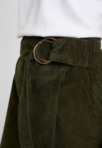 Primer plano de unos pantalones de pana verde oliva con un cinturón de tela a juego asegurado por dos anillas en D de latón, llevados con una blusa blanca metida por dentro.