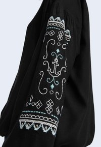 Blusa nera a maniche lunghe con complessi ricami bianchi e turchesi sulle maniche, caratterizzata da motivi decorativi e un motivo ancorato.