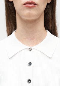 Κοντινό πλάνο του κατώτερου μέρους του προσώπου και του λαιμού ενός ατόμου με μια οριζόντια ουλή πάνω από ένα λευκό πουκάμισο με ασημένια κουμπιά.