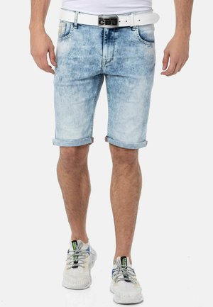 Mann trägt hellblaue, hochgekrempelte Jeansshorts, einen weißen Gürtel mit CBJ-Schnalle und weiße Sneaker mit schwarzen und grünen Tag-Details.