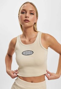 Beige ribbet crop top har en rund halsudskæring og et grafisk logo-patch i sort i midten. Materialet har en blød tekstur og en tætsiddende pasform.