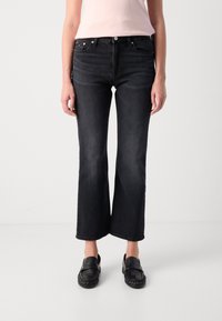 Kvinne iført svart bootcut-jeans, svarte loafers og en lys rosa ermeløs topp, stående mot en enkel lys bakgrunn.