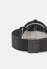 Svart rustfritt stål mesh klokke med rund kasse, minimalistisk design og polert lås. Har en gjennomsiktig bakkasse.