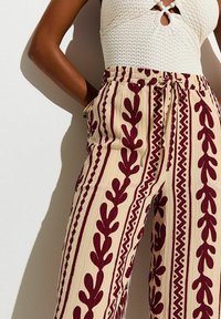 Mujer con pantalones crema de talle alto con patrones de hojas y zigzag en color burdeos, combinados con una camiseta sin mangas blanca texturizada.
