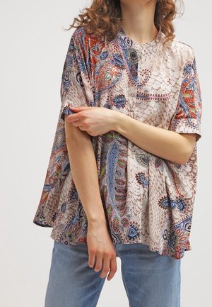 Femme portant un chemisier ample à motif cachemire avec des détails rouges, bleus et verts, associé à un jean bleu clair, posant les bras croisés.