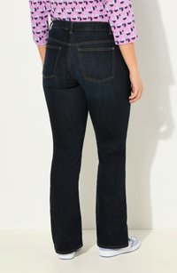 Mörkblå utsvängda jeans med midjeband, bakfickor och kontrastsömmar. Kombineras med vita slip-on skor mot en neutral bakgrund.