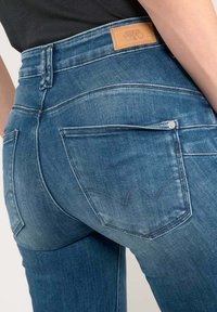 Jean en denim bleu foncé avec une taille haute, des coutures subtiles et deux poches arrière présentant un design signature sur le tissu.