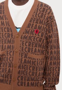 Καφέ πλεχτό καπιτονέ με επαναλαμβανόμενο σχέδιο κειμένου "ΠΑΓΩΤΟ", δύο μπροστινές τσέπες και ένα κόκκινο κεντημένο λογότυπο με γραφικό σχέδιο.