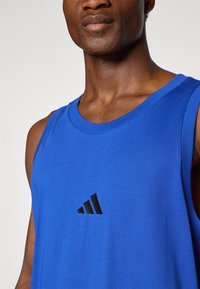 Modra bombažna majica brez rokavov s krožnim izrezom in črnim Adidasovim logotipom. Tkanina ima gladko teksturo in sproščen kroj.