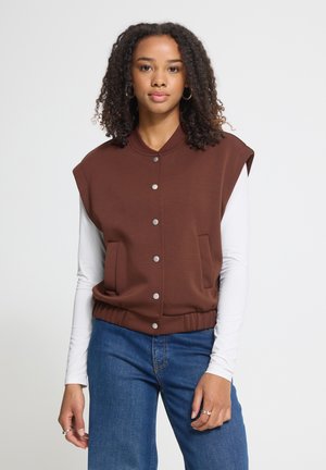 Jeune femme aux cheveux bouclés portant un gilet marron sans manches boutonné sur une chemise blanche à manches longues et un jean bleu, face à l'avant.