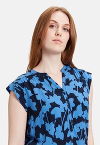 Kortærmet bluse med mørkeblåt baggrund og lyseblå blomsterprint; har V-udskæring og knapdetalje.