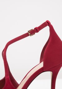 Röd mockaskinn högklackad sko med ankelrem och gyllene spänne, vinklad sidovy på vit bakgrund.