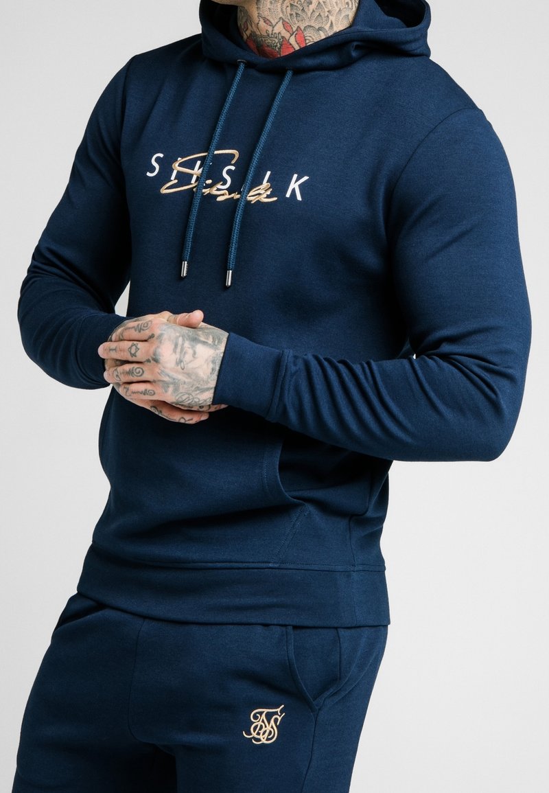 Homme tatoué portant un sweat à capuche bleu marine et un pantalon de survêtement assorti avec des logos brodés beige, mains jointes à la taille.