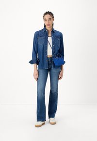 Donna con capelli intrecciati che indossa una camicia di jeans sopra un crop top bianco, jeans blu, sneakers bianche e una piccola borsa a tracolla blu.