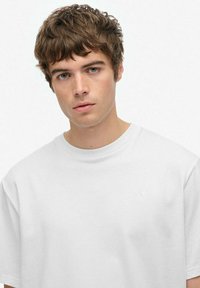 Ung mand med kort, krøllet brunt hår og blå øjne, iført en enkel hvid T-shirt med rund hals, vendt mod kameraet med en neutral udtryksform.