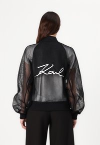 Black mesh bomber jacket with long, sheer sleeves, ribbed cuffs, and waistband. Features large white "Karl" signature on the back.