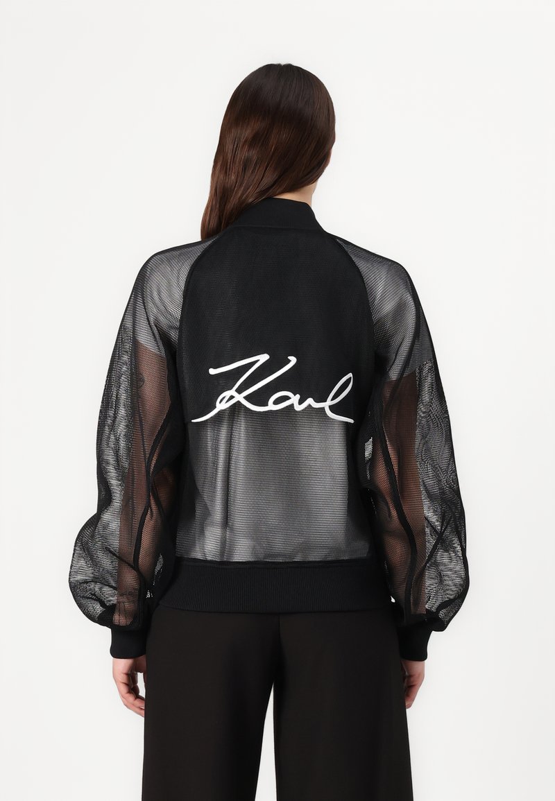 Black mesh bomber jacket with long, sheer sleeves, ribbed cuffs, and waistband. Features large white "Karl" signature on the back.