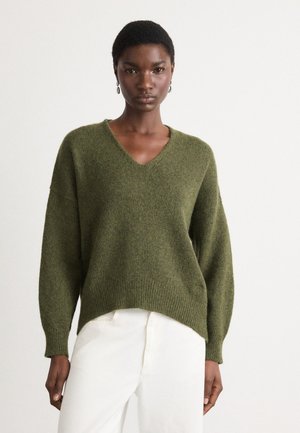 Oliven grønn, oversized strikkegenser med V-hals og senkede skuldre. Ribbede mansjetter og bunnkant, kombinert med hvite bukser.