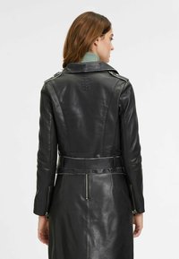 Schwarzer Leder-Trenchcoat mit tailliertem Schnitt, Schulterklappen und Reißverschlüssen an den Ärmelbündchen. Die glatte Textur und das maßgeschneiderte Design betonen die Silhouette.