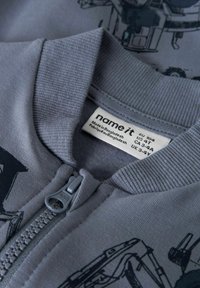 Grå zip-up jakke med ribbet krave, der har et grafisk print og et label med størrelses- og plejeinformation. Glat stofstruktur.