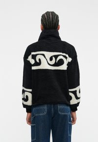Fekete és fehér fleece pulóver magas gallérral, forgó mintákkal az ujjakon. Texturált, laza szabású design.